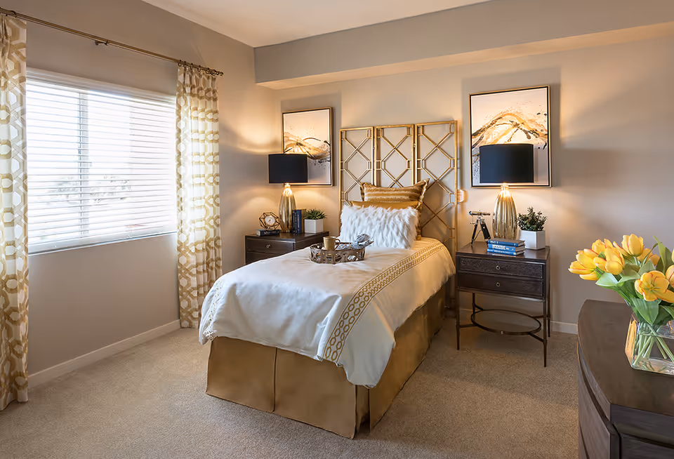 A cozy bedroom with a single bed featuring a decorative gold and white headboard, white and gold bedding, and a fluffy white pillow. There are two dark wooden nightstands on either side of the bed, each with a gold lamp and decorative items. The room has beige carpet, a window with patterned gold and white curtains, and two abstract paintings above the bed. A vase with yellow tulips is on a dresser to the right.