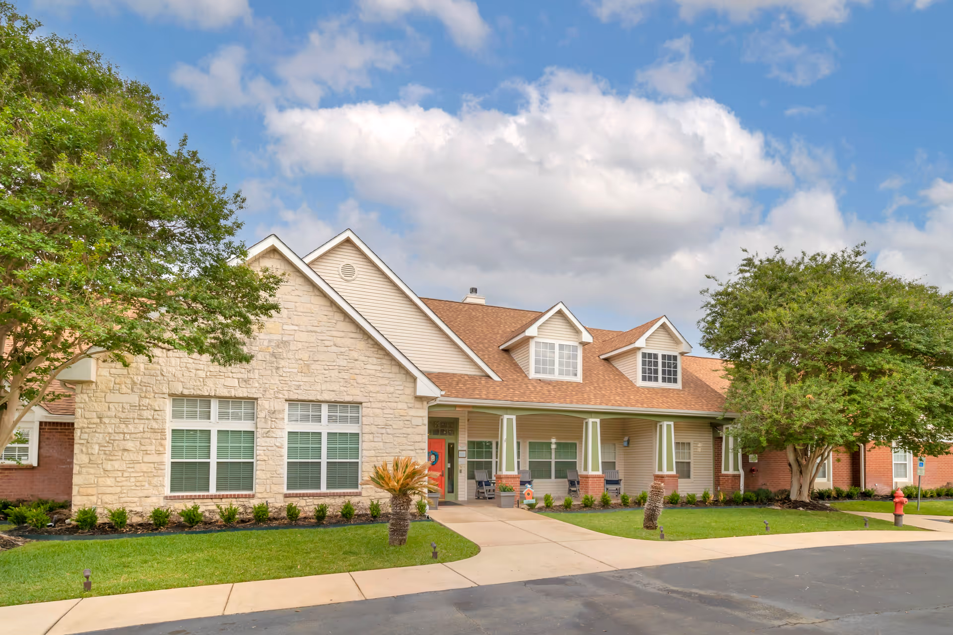 Exterior view of a single-story senior living facility building with a stone and brick facade, multiple windows, a covered porch with columns, green lawn, trees, and a partly cloudy sky.