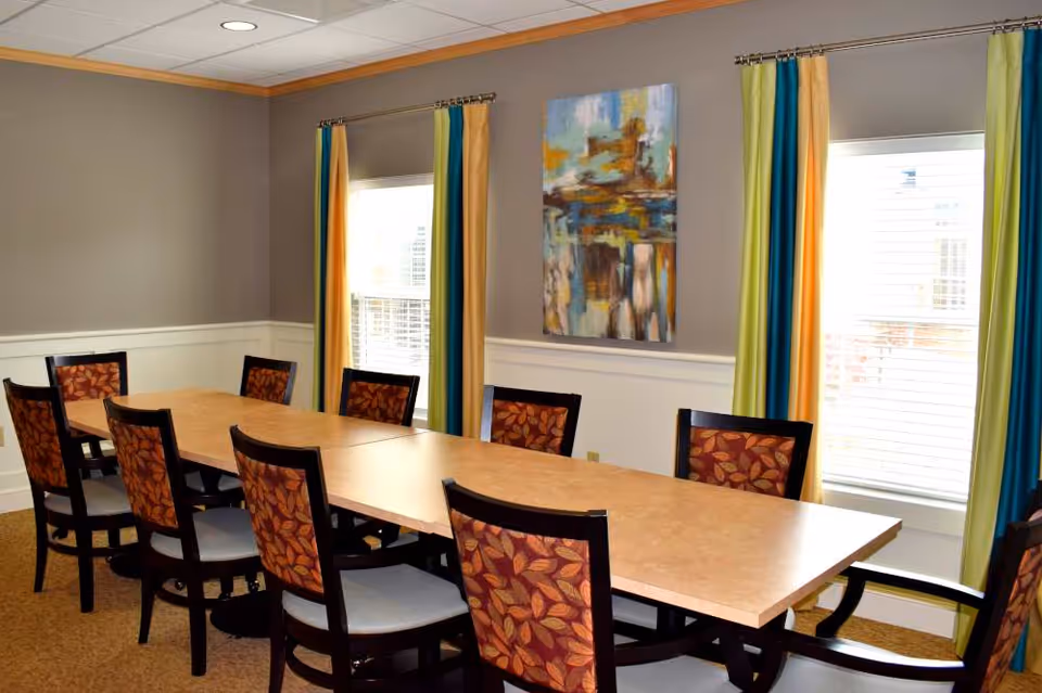 A dining room with a long rectangular wooden table surrounded by ten chairs with patterned upholstery. The room has two windows with colorful curtains in shades of yellow, green, and blue. A modern abstract painting hangs on the wall between the windows. The walls are painted gray with white wainscoting, and the ceiling has recessed lighting.