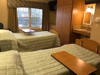 A nursing facility bedroom with two single beds covered in green bedspreads. Each bed has a wooden overbed table. There is a large window with curtains, a wooden wardrobe, a wall-mounted TV, and a small sink with a mirror and light above it.