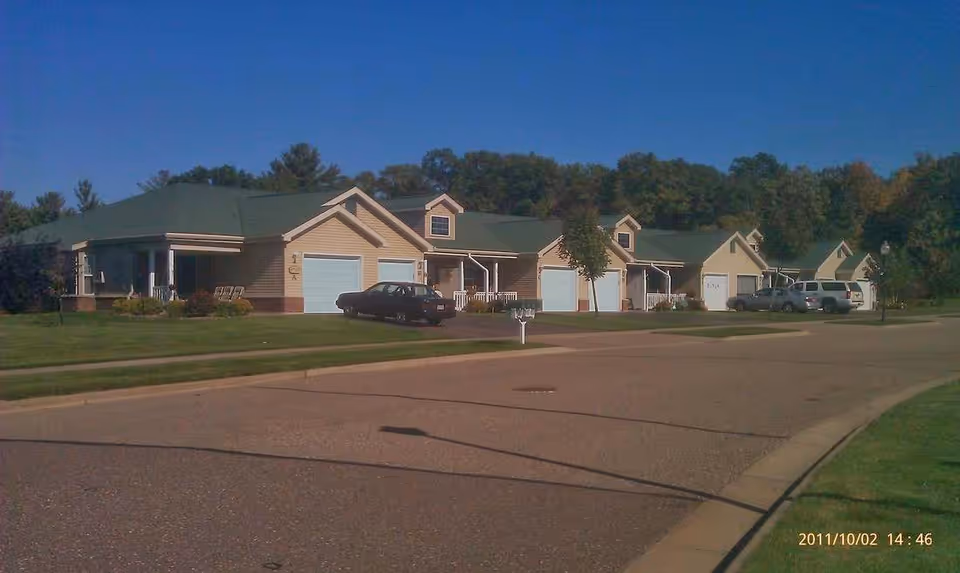 A row of single-story residential cottages with green roofs and beige siding, each with a garage and driveway. Several cars are parked in the driveways. The street curves in front of the cottages, and there are trees and greenery in the background under a clear blue sky.