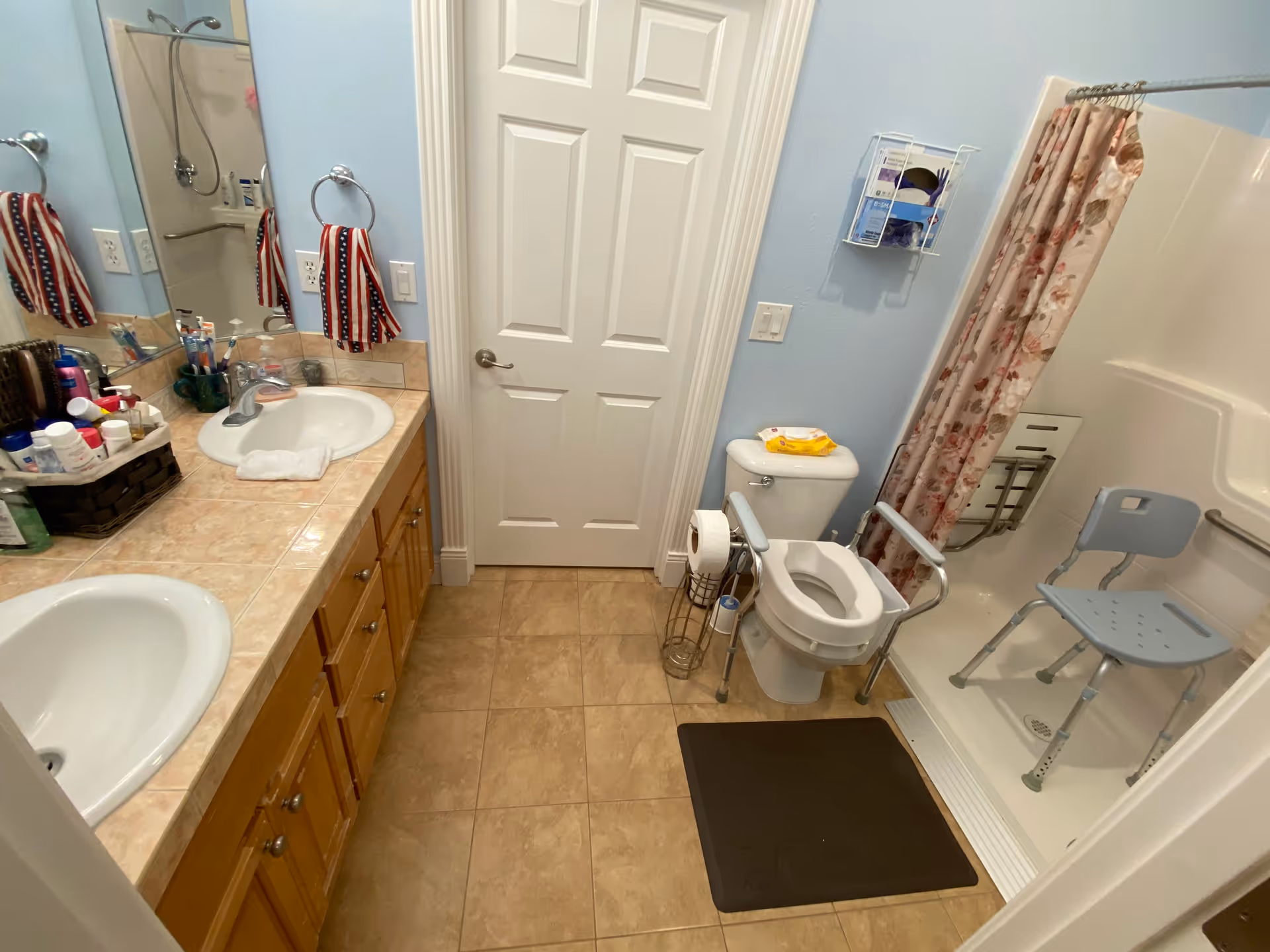 A bathroom with two sinks and a large mirror on the left side, a toilet with safety rails in the center, and a shower area with a floral shower curtain and a blue shower chair on the right. The floor is tiled, and there are various toiletries on the countertop.