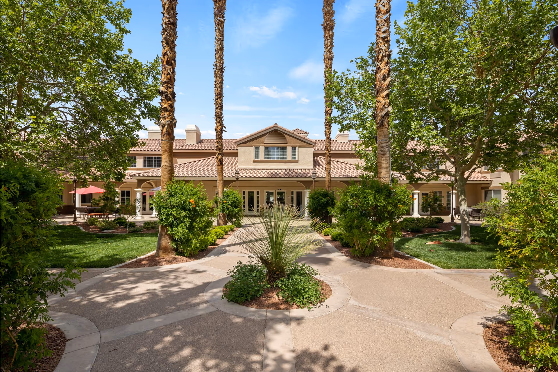 Exterior view of Oakmont of Las Vegas facility showing a pathway lined with palm trees and greenery leading to the building entrance. The building has a tiled roof and multiple windows, with a clear blue sky above.