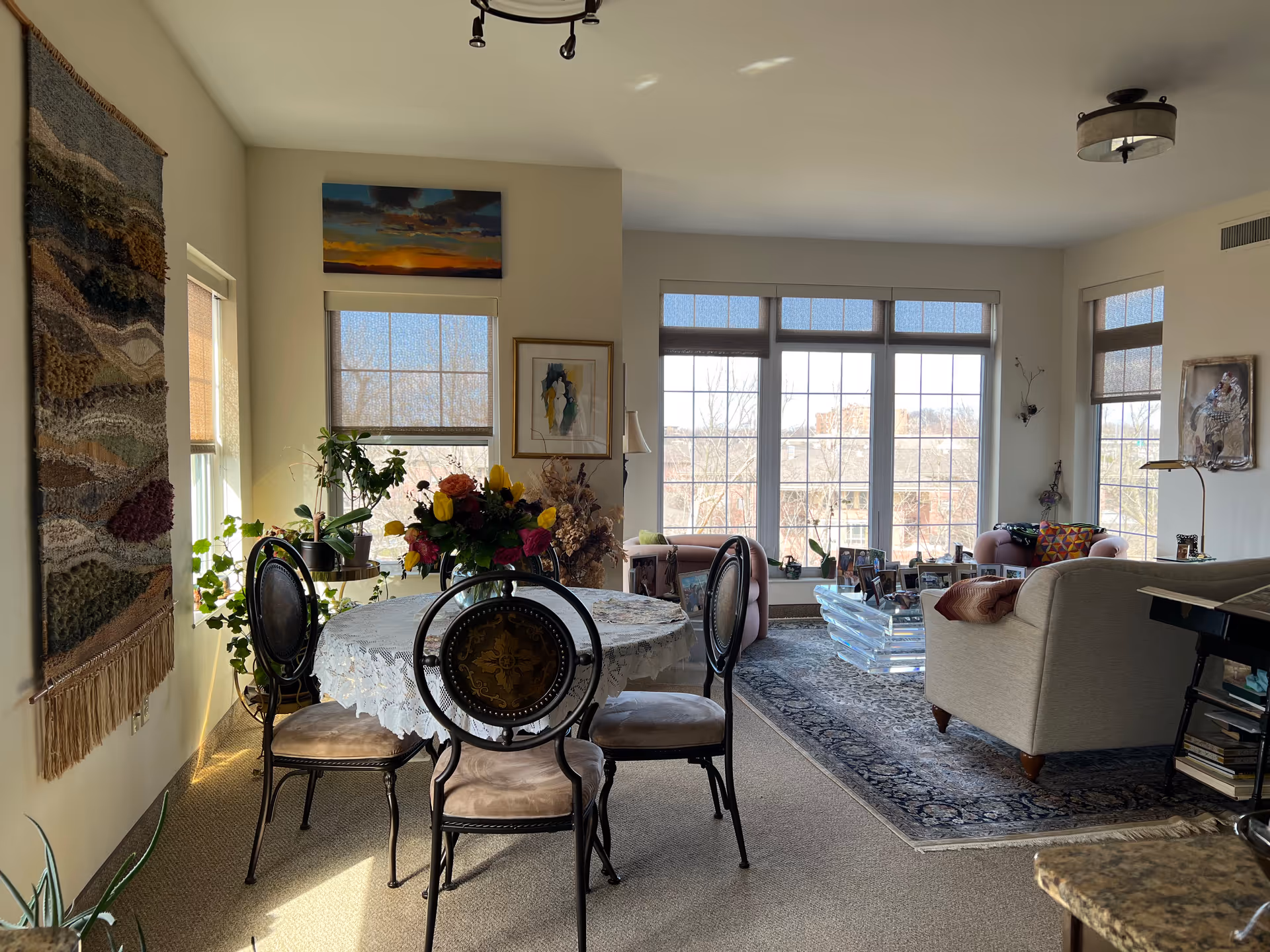 A bright and cozy living and dining area with large windows letting in natural light. The dining area features a round table covered with a lace tablecloth and four ornate chairs. There are various plants and a colorful floral centerpiece on the table. The living area has a beige sofa, two pink armchairs, a glass coffee table with framed photos, and a patterned rug. Wall art and a woven tapestry decorate the walls.