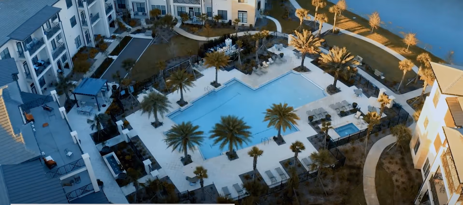 Aerial view of an outdoor swimming pool area surrounded by palm trees, lounge chairs, and tables within a residential or assisted living community. The pool area is enclosed by a fence and adjacent to multi-story buildings with balconies. There are paved walkways and landscaped green spaces around the pool.