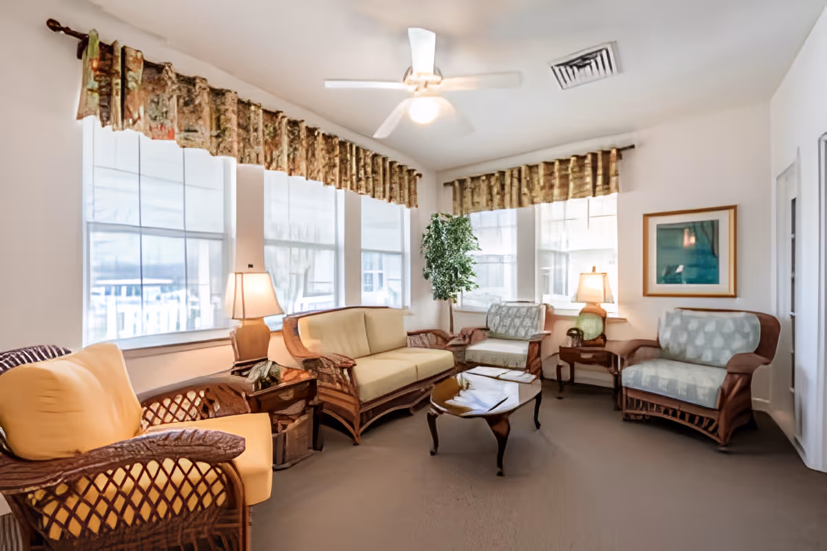 A cozy living room with wicker furniture including a sofa, three armchairs with cushions, two side tables with lamps, a coffee table, a potted plant, and large windows with sheer curtains and valances. The ceiling has a white fan with a light.