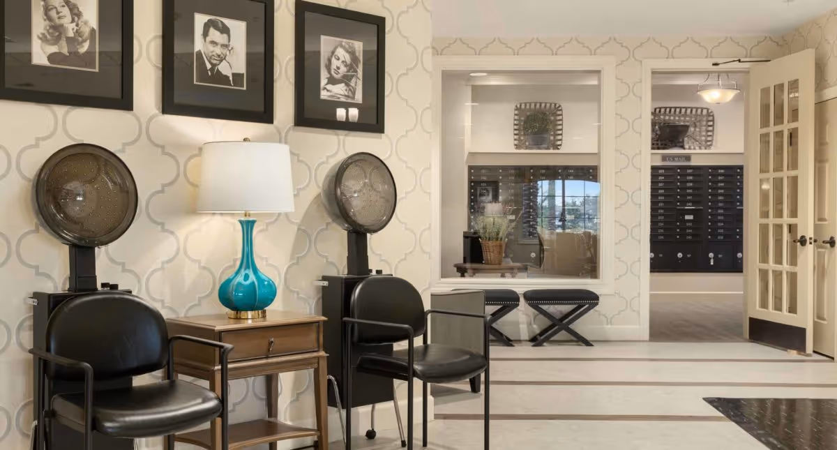 Interior view of a senior living facility hair salon area with two black salon chairs and hair dryers, a wooden side table with a turquoise lamp, and three black and white framed portraits on the wall. In the background, there is a window looking into a room with benches and a wall of mailboxes.