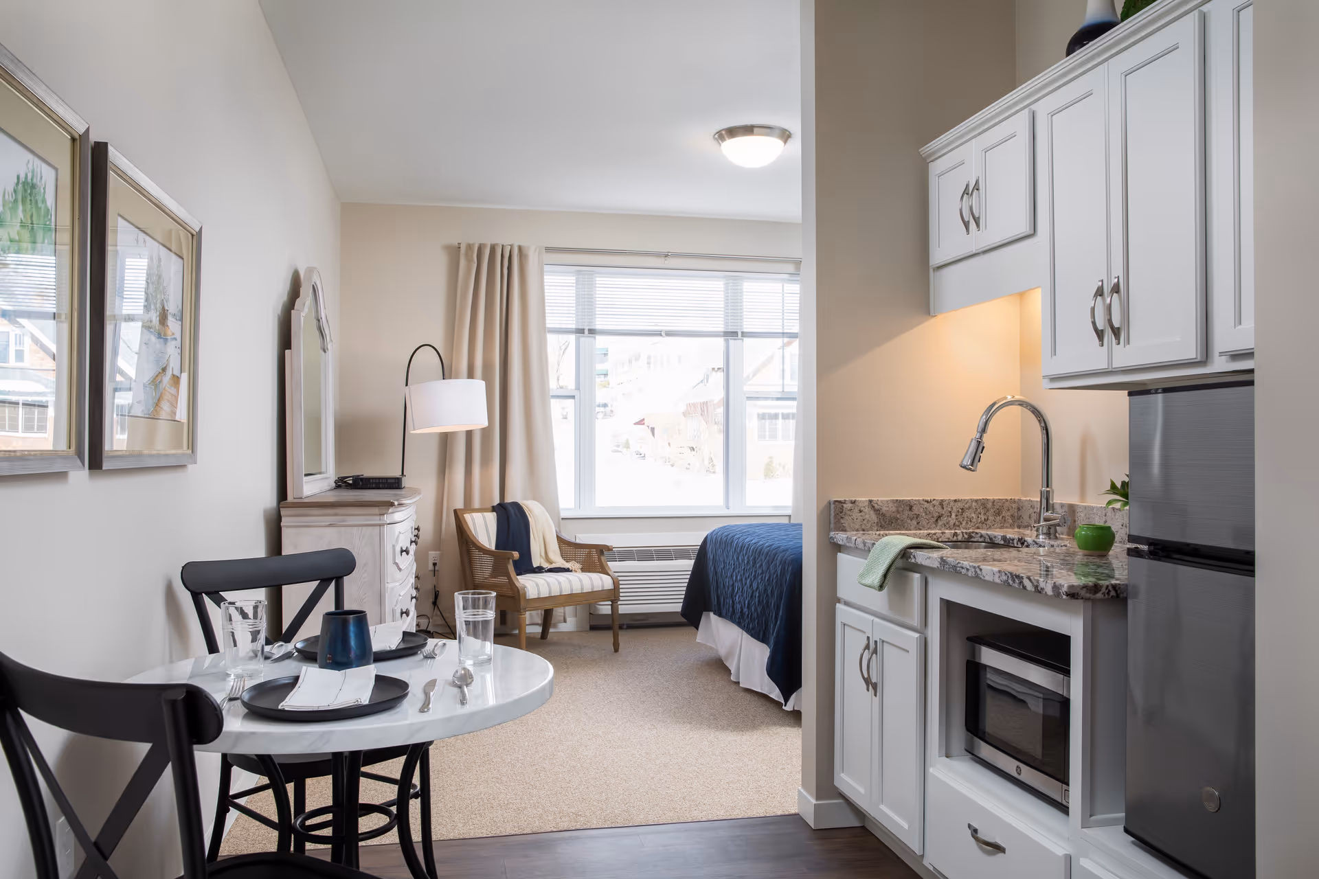 A cozy studio apartment featuring a small kitchenette with white cabinets, a granite countertop, a sink, microwave, and mini refrigerator. Adjacent to the kitchenette is a round dining table set for two with black chairs. In the background, there is a sitting area with a cushioned chair, a floor lamp, a dresser with a mirror, and a bed with a blue quilt near a large window with beige curtains letting in natural light.