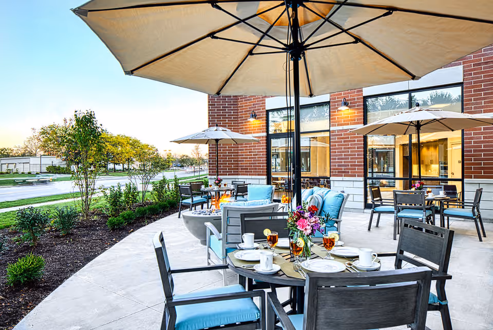 Outdoor patio area at The Sheridan at Park Ridge with tables and chairs under large beige umbrellas. The tables are set with plates, cups, glasses of iced tea, and a vase of flowers. The patio is adjacent to a brick building with large windows, and there are plants and trees along the edge of the patio near the street.