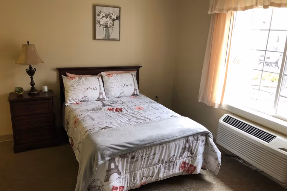A bedroom with a double bed featuring Paris-themed bedding, a wooden nightstand with a lamp and small decorative items, a framed floral picture on the wall, and a large window with curtains letting in natural light. There is also a heating/cooling unit below the window.