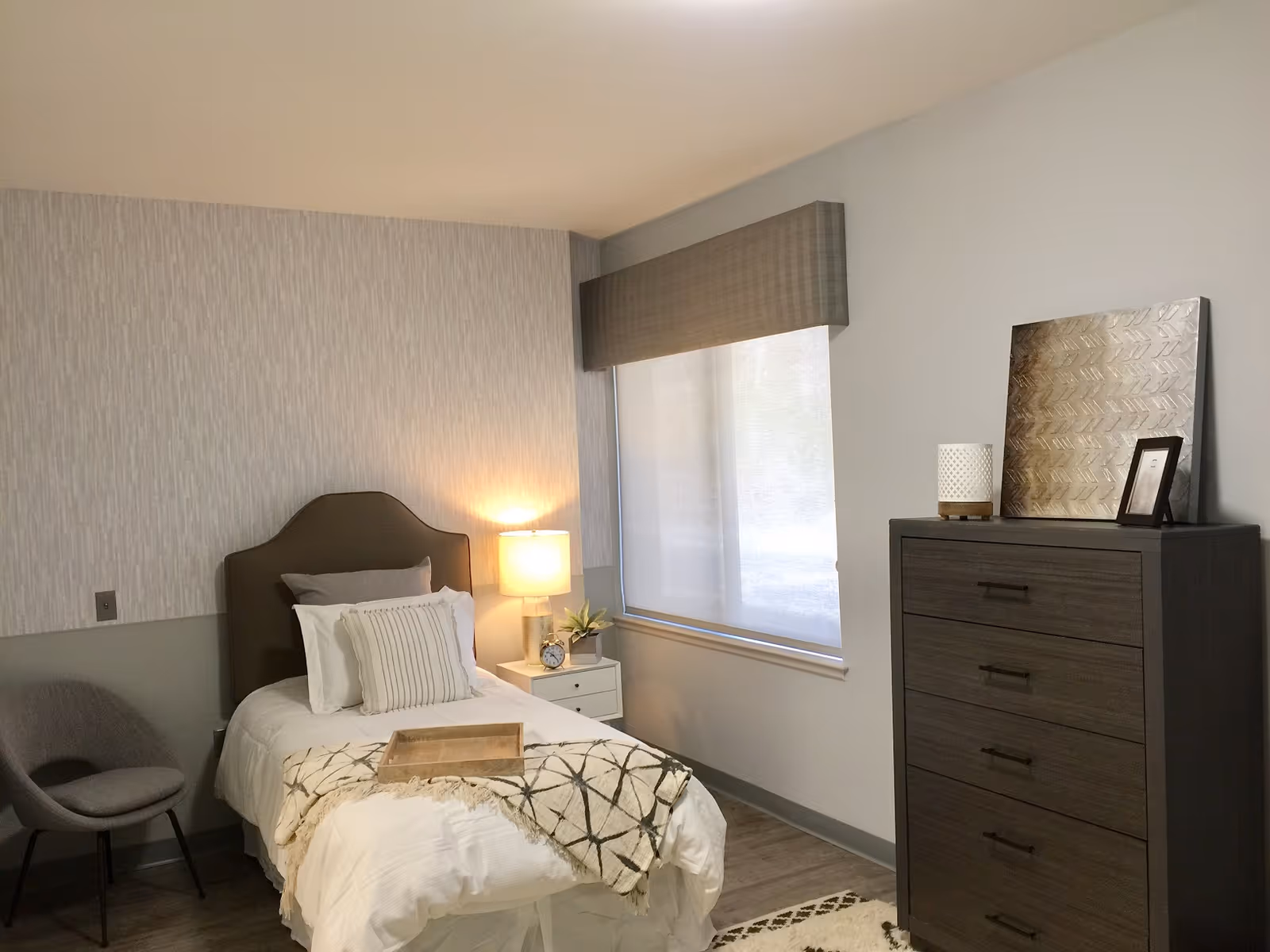 A neatly arranged bedroom with a single bed featuring white bedding and decorative pillows. A wooden tray is placed on the bed. Next to the bed is a small white nightstand with a lamp, a clock, and a small plant. A gray upholstered chair is positioned in the corner. A large window with a gray valance and a roller shade lets in natural light. On the right side of the room is a dark wooden dresser with four drawers, topped with decorative items including a framed picture and a textured artwork. The walls are painted in light neutral tones, and the floor has a wood-like finish.