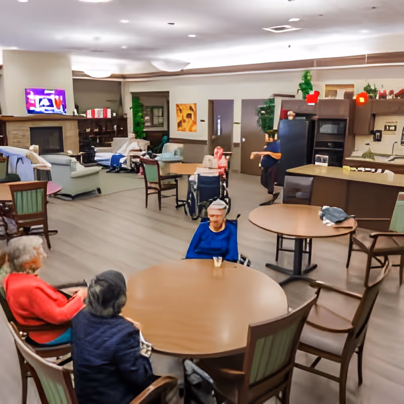 A common area in a senior living facility with several elderly residents seated around round tables. The room has a cozy atmosphere with a fireplace, a television mounted above it, and a kitchen area in the background. The floor is wooden, and there are various chairs and wheelchairs present.