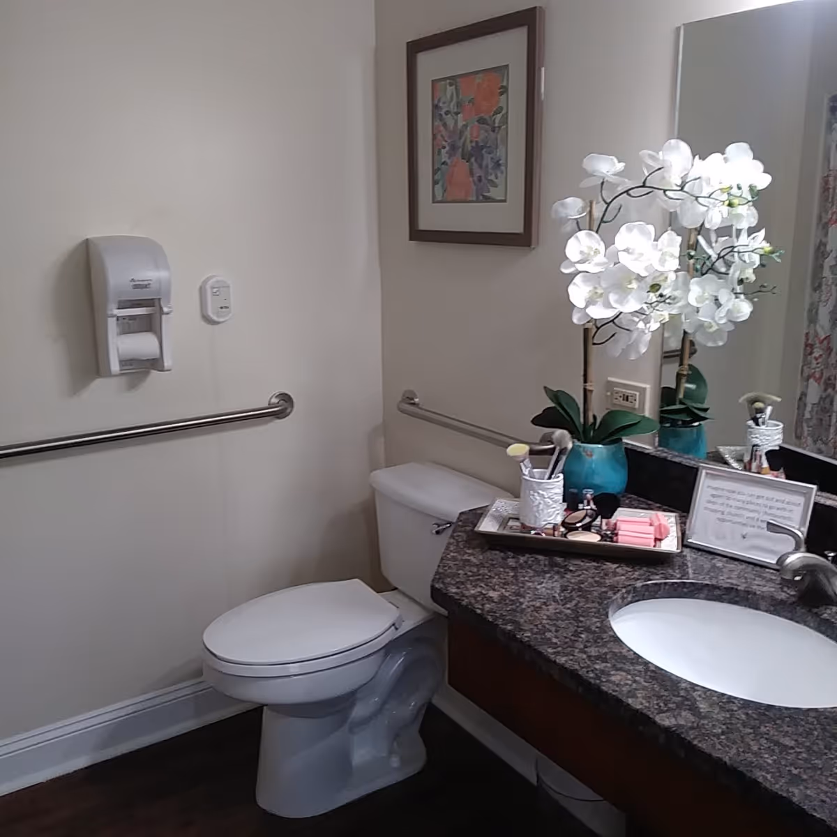 A bathroom with a white toilet, a granite countertop with a sink, a vase with white orchids, makeup items on a tray, a framed floral picture on the wall, and safety grab bars on the walls.