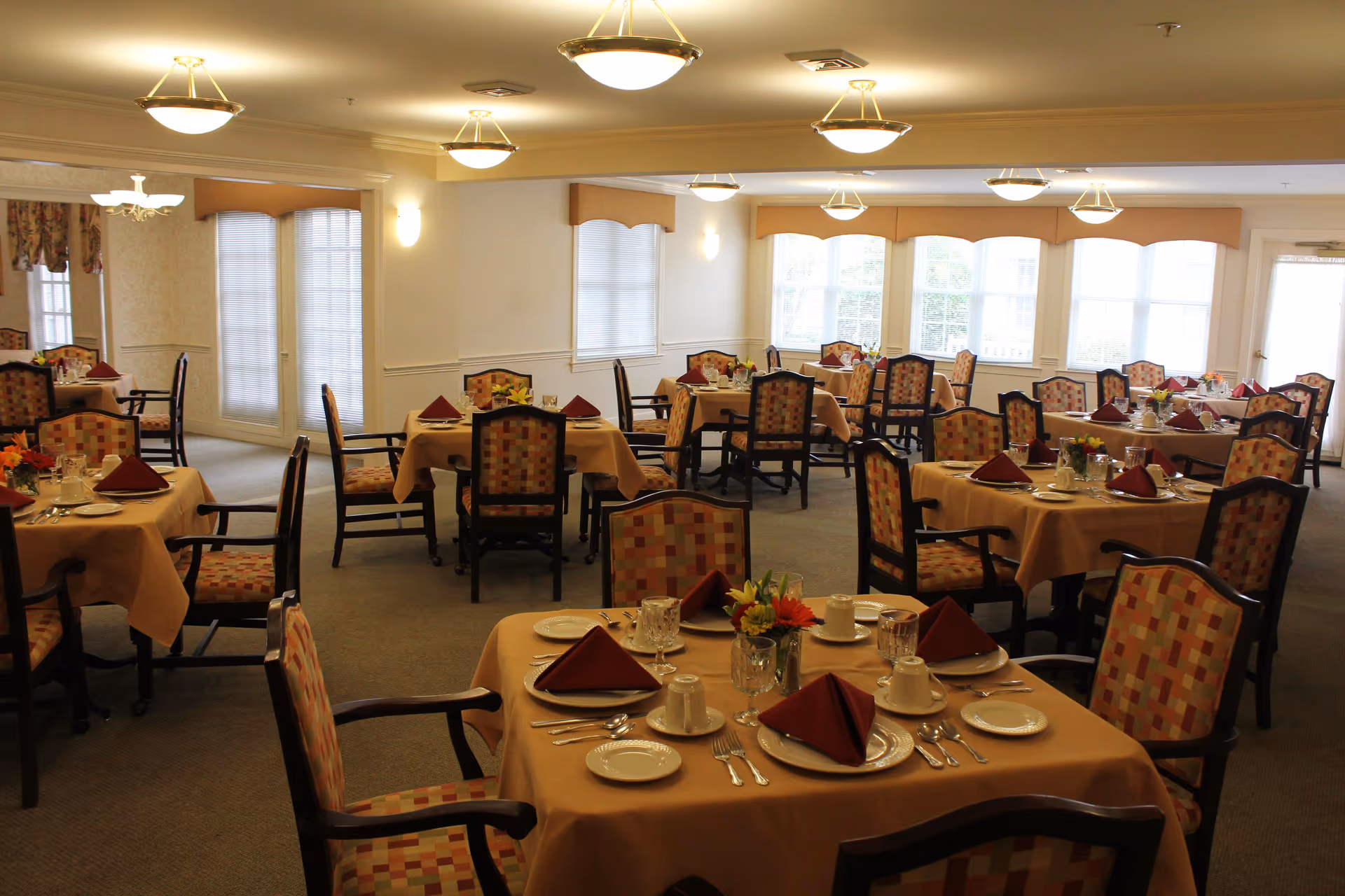 A dining room in an assisted living facility with multiple tables covered in beige tablecloths, each set with plates, cups, silverware, and folded maroon napkins. The chairs have patterned upholstery, and the room is well-lit with ceiling lights and large windows allowing natural light.