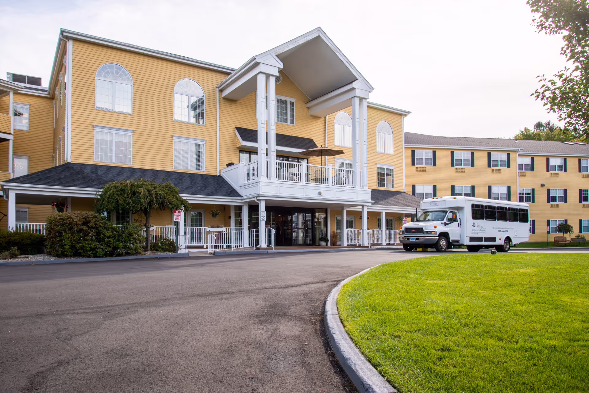 Yellow three-story senior living building with a covered main entrance and a white shuttle bus parked in front.