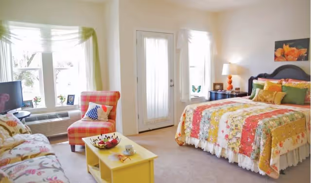 A bright and cozy bedroom featuring a bed with a colorful floral quilt and multiple pillows, a nightstand with a lamp and decorative items, a plaid armchair with a star-patterned pillow, a floral sofa, a yellow coffee table with a cup and a bowl, and large windows with sheer curtains letting in natural light.