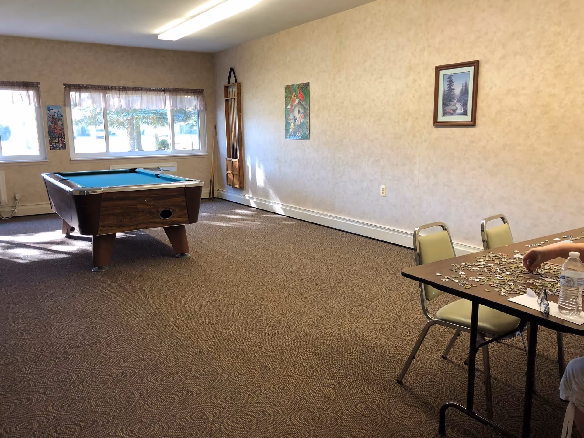 A room with a pool table near windows letting in natural light, a wall-mounted rack holding pool cues, and a table with chairs where a person is working on a jigsaw puzzle. The walls have framed pictures and the floor is carpeted.