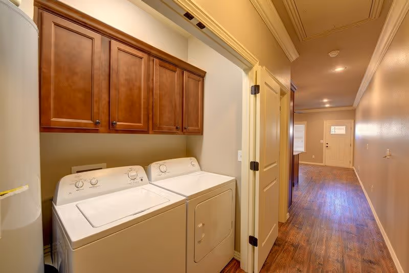 Interior hallway leading to a laundry nook with a washer and dryer beneath wooden cabinets and wood-look flooring.