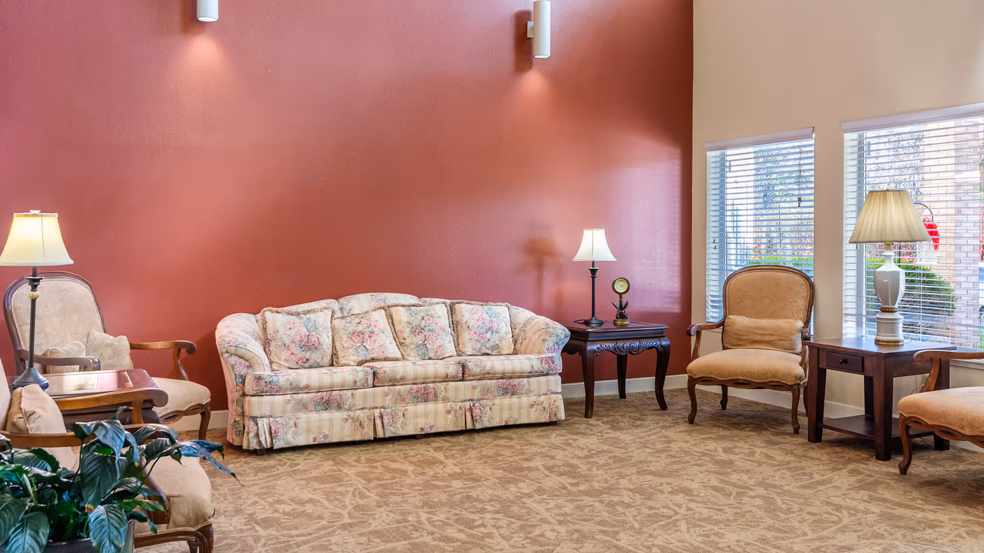 A cozy living room area with a floral patterned sofa, two beige armchairs, wooden side tables with lamps, and large windows with blinds letting in natural light. The walls are painted in a warm red and beige color, and the floor is carpeted with a patterned design.