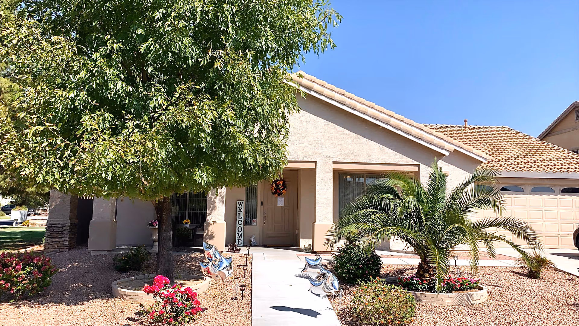 Front exterior view of a single-story house with a tiled roof, a large tree on the left, a palm plant on the right, and a concrete walkway leading to the front door. There are decorative star-shaped balloons and a vertical 'WELCOME' sign near the entrance. The landscaping includes rocks and flowering plants under a clear blue sky.