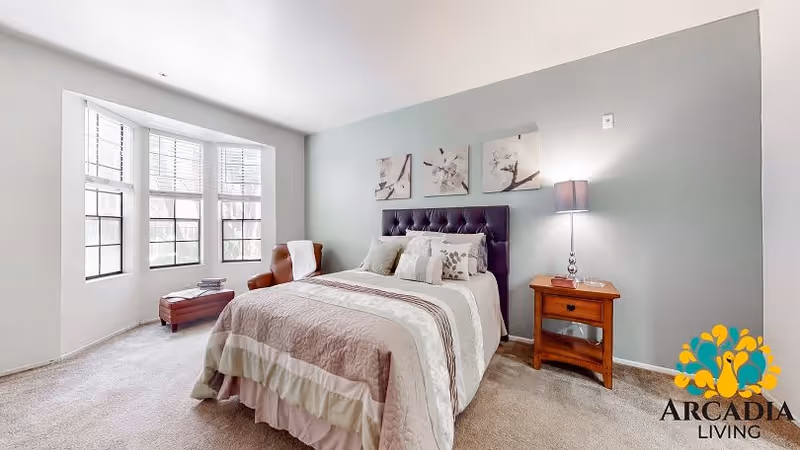 Well-lit furnished bedroom with a bed, decorative pillows, a nightstand and lamp, an armchair by a bay window, and an Arcadia Living logo.