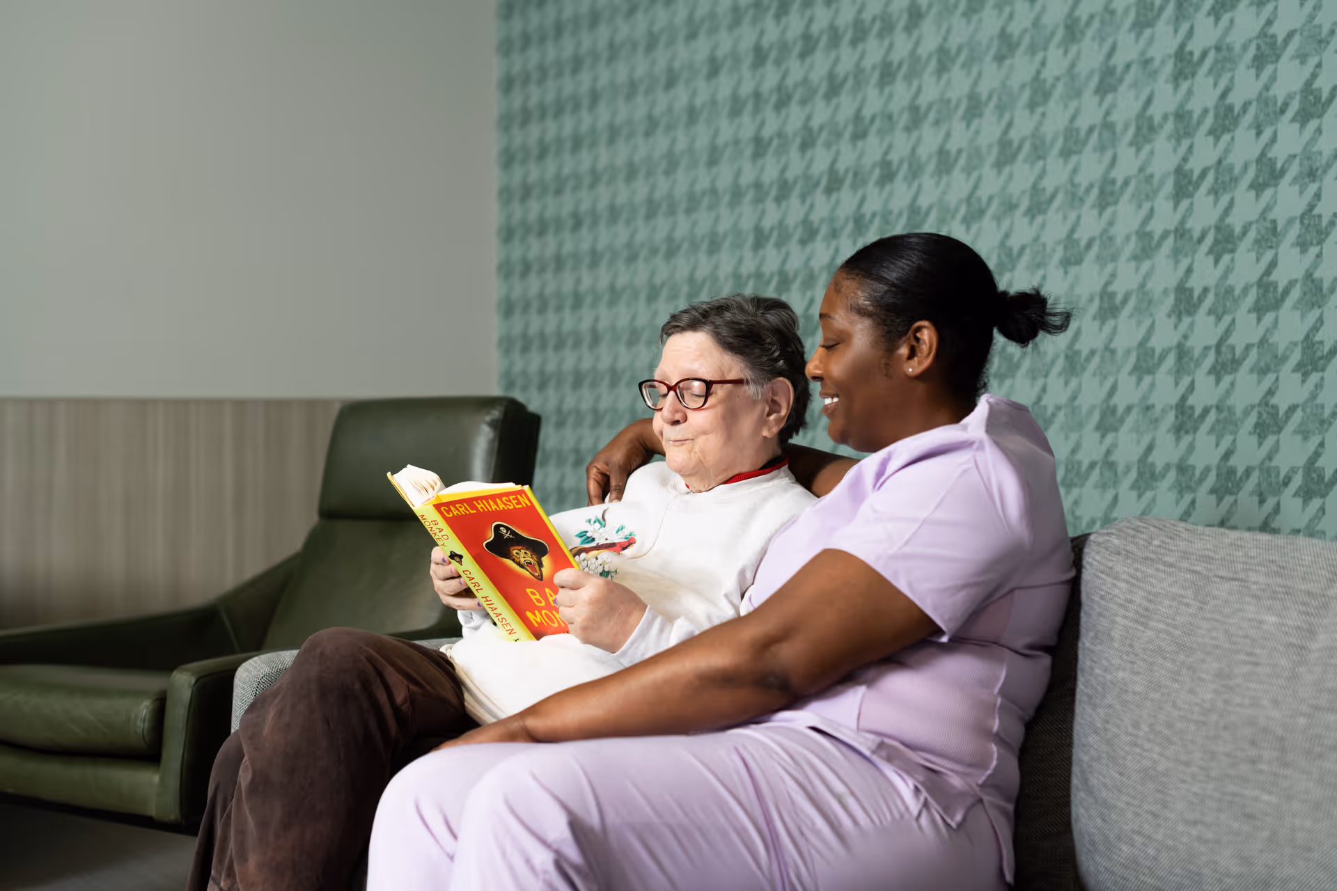 An elderly woman wearing glasses and a white sweater is sitting on a couch reading a book titled 'Bad Monkey' by Carl Hiaasen. Next to her, a caregiver in a light purple uniform is sitting closely, smiling and looking at the book. The background features a green patterned wall and a green armchair.