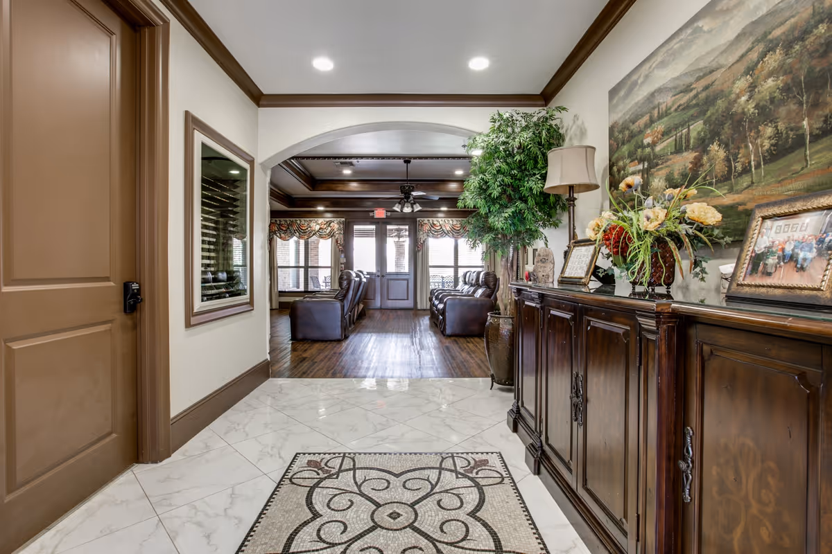 Interior view of a senior living facility hallway with a decorative tiled floor mat, wooden cabinet with floral arrangement and framed pictures, a large painting on the wall, and a potted plant. The hallway leads to a common area with leather armchairs and large windows with patterned valances.