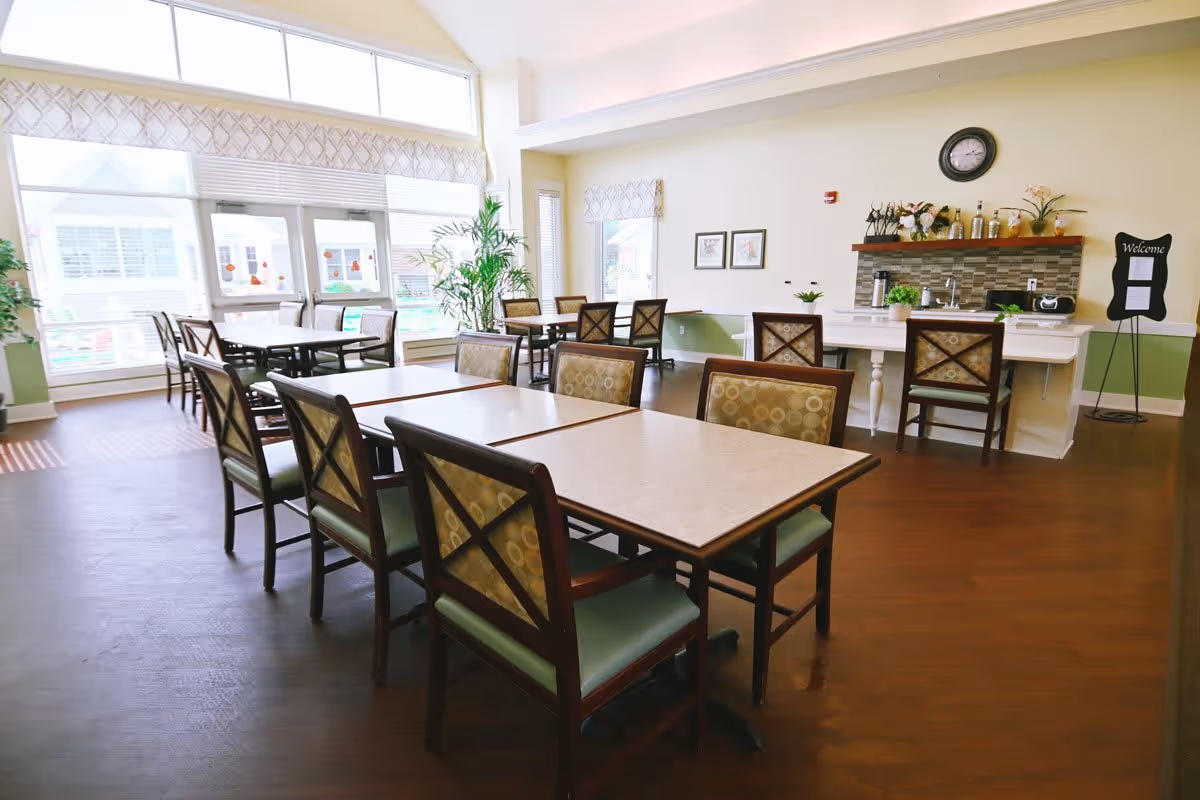 A bright dining room with multiple tables and chairs arranged neatly. Large windows allow natural light to fill the room. There is a counter area with chairs, decorative plants, and a clock on the wall. A welcome sign is displayed on a stand near the counter.