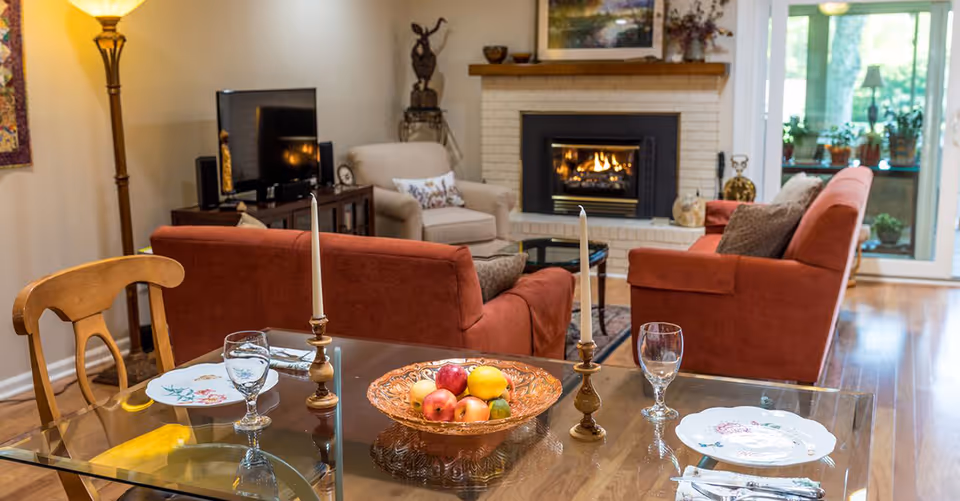 Open-plan interior showing a glass dining table set with plates and glassware in the foreground and red sofas with a lit fireplace in the living area behind it.