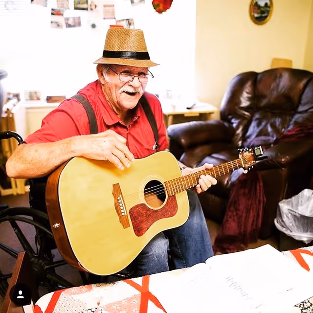 An elderly man wearing a straw hat, glasses, and a red shirt with suspenders is sitting in a wheelchair and playing an acoustic guitar in a cozy living room. Behind him, there is a brown leather armchair and a small table with a lamp. The room has warm yellow walls with some decorations and photos pinned on a board.