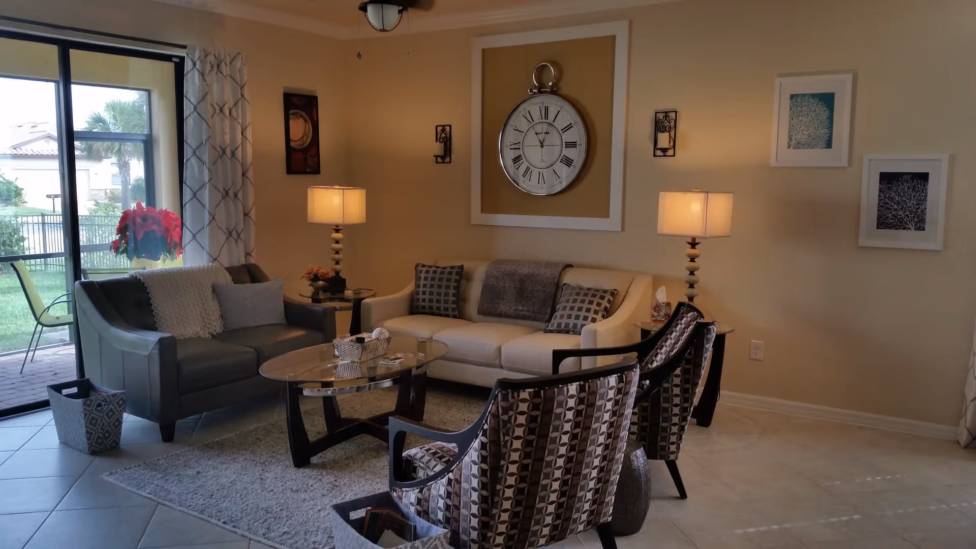 A cozy living room with a beige sofa, two patterned armchairs, a dark gray loveseat, and a glass coffee table on a light gray rug. Two table lamps with beige shades are on either side of the sofa. A large decorative clock is mounted on the wall above the sofa, flanked by two candle sconces. There are framed artworks on the walls and a sliding glass door leading to an outdoor patio with greenery visible outside.