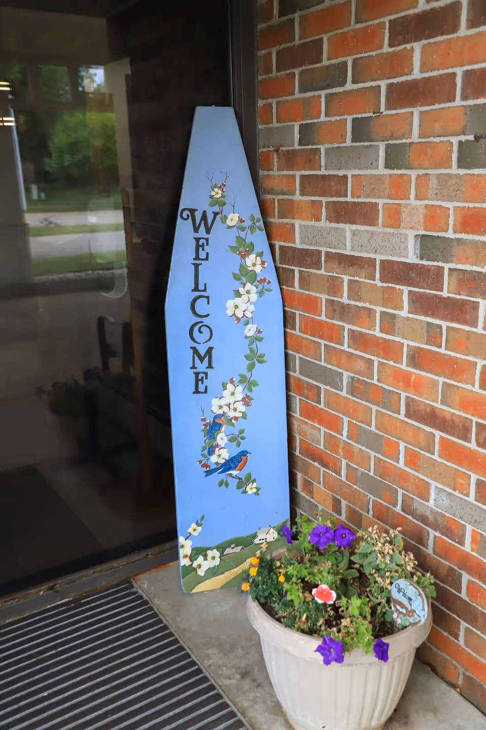 A blue wooden welcome sign decorated with painted white flowers and two bluebirds, leaning against a brick wall next to a white planter filled with purple and yellow flowers and green foliage, placed near a glass door entrance.