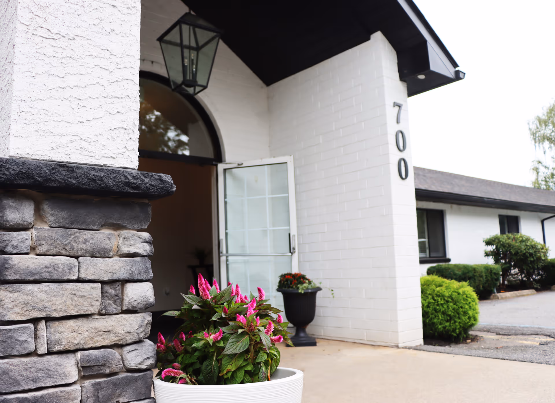 Entrance of a building with white brick walls and the number 700 displayed vertically. There is a stone pillar on the left side and a white door that is open. A white planter with pink flowers is in the foreground, and a black lantern hangs above the entrance.