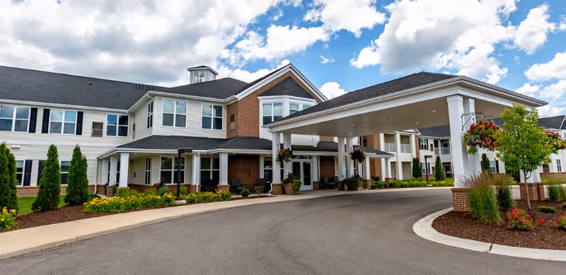Exterior view of a two-story assisted living facility with a covered entrance driveway, landscaped garden beds with flowers and shrubs, and a partly cloudy blue sky.