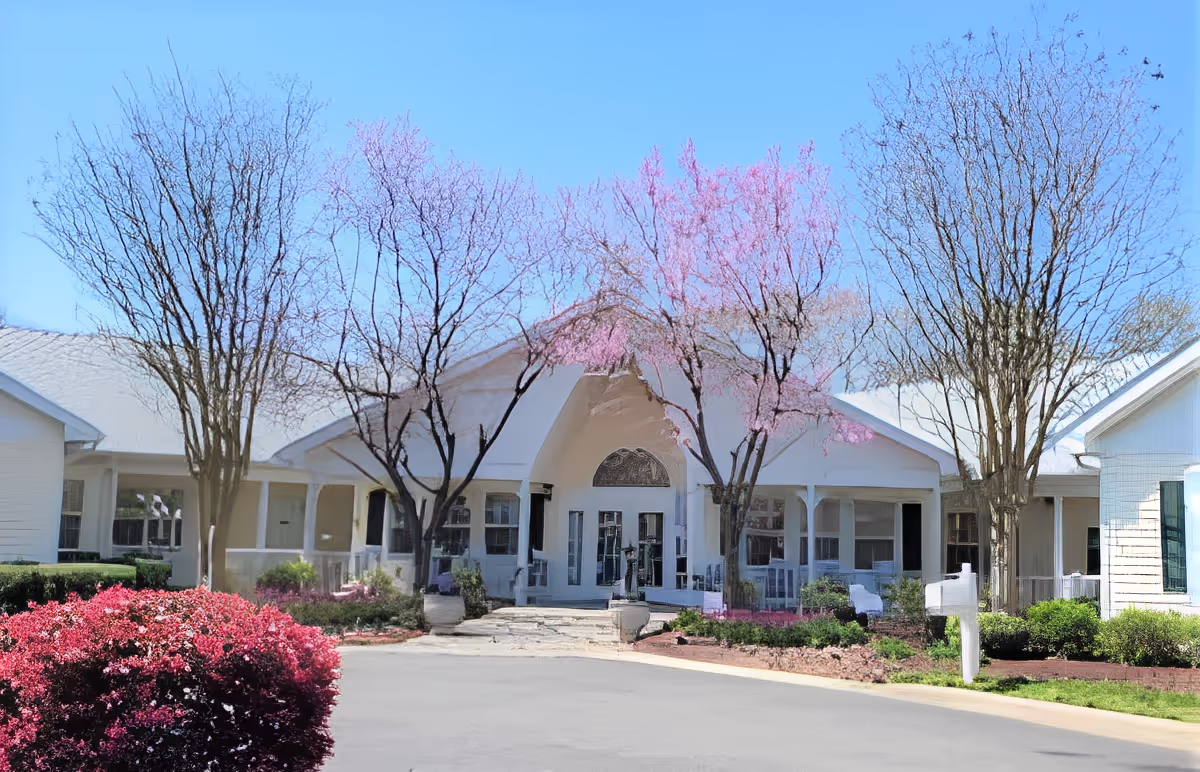 White single-story building entrance with an arched portico, blooming pink trees, and landscaped driveway.