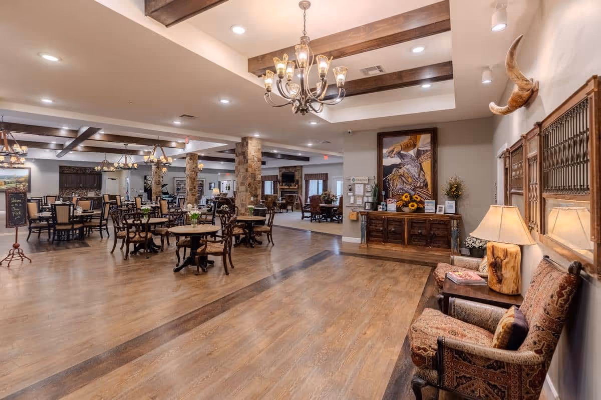 Spacious dining area in an assisted living facility with multiple wooden tables and chairs arranged neatly. The room features wooden beams on the ceiling, stone pillars, chandeliers, and warm lighting. A cozy seating area with upholstered chairs and a lamp is visible on the right side, along with decorative wall art and a mounted horn.