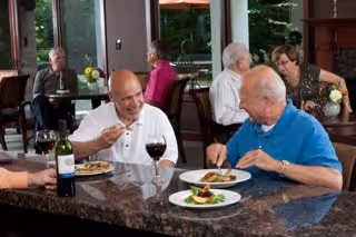 Two elderly men sitting at a dining table in a communal dining area, enjoying a meal and wine. Other seniors are seated at tables in the background near large windows with a view of greenery outside.