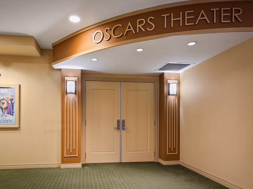 Entrance to Oscars Theater with double beige doors, wall sconces on either side, and a sign above the doors reading 'OSCARS THEATER'. A framed Frozen movie poster is visible on the left wall. The walls are painted beige and the floor is carpeted in green with a subtle pattern.