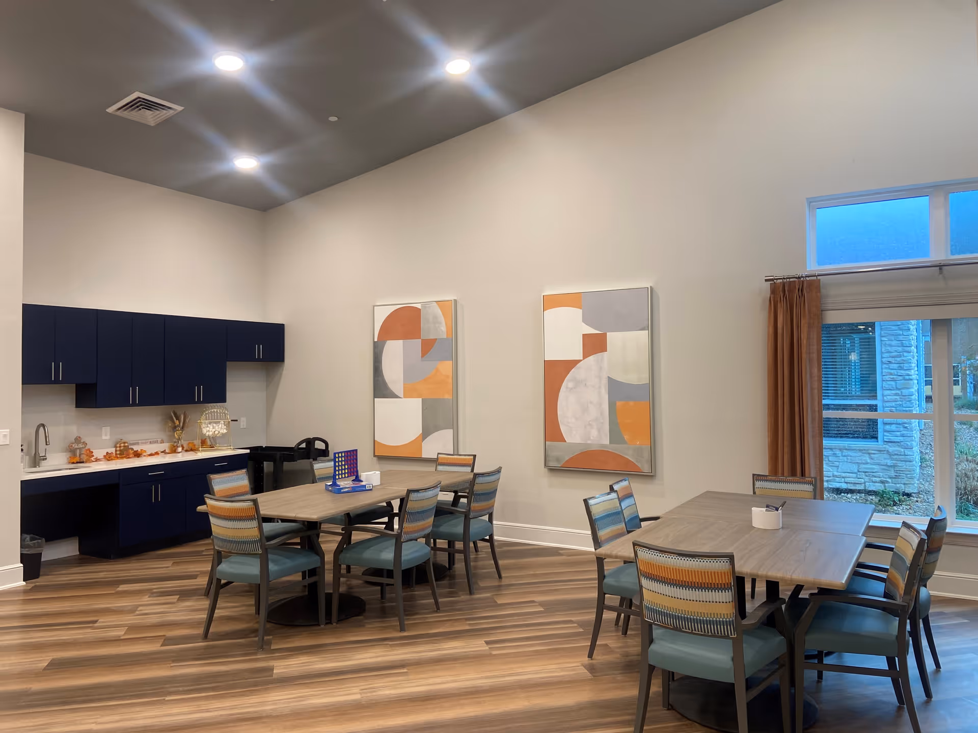 Interior room with two wooden tables surrounded by chairs with blue cushions and multicolored backs. The room has wood flooring, a set of dark blue cabinets with a countertop and sink on the left wall, and two abstract art paintings with orange, gray, and white shapes on the wall. A large window with brown curtains shows an outdoor view.