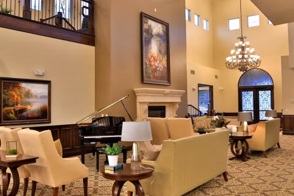 A spacious and elegant living room area with high ceilings, a grand chandelier, and large windows. The room features beige sofas and armchairs arranged around wooden side tables with lamps and plants. A black grand piano is positioned near a fireplace with a large framed painting above it. Another framed painting hangs on the wall to the left, and a decorative double door with glass panels is visible in the background.