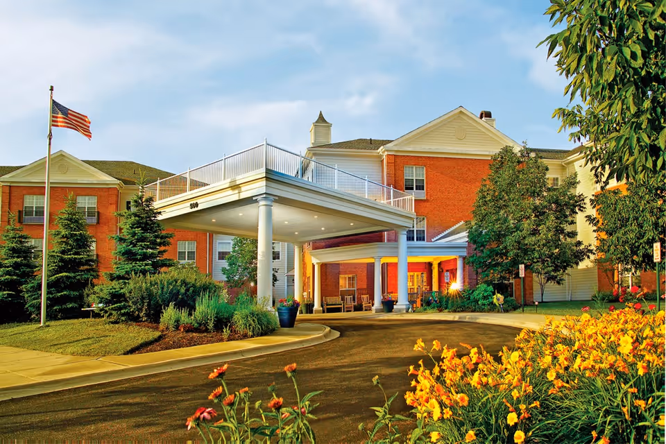 Exterior view of Belmont Village Senior Living Buffalo Grove showing a large brick and siding building with a covered entrance supported by white columns. There is a driveway leading up to the entrance, landscaped greenery, colorful flowers in the foreground, and an American flag on a flagpole to the left.