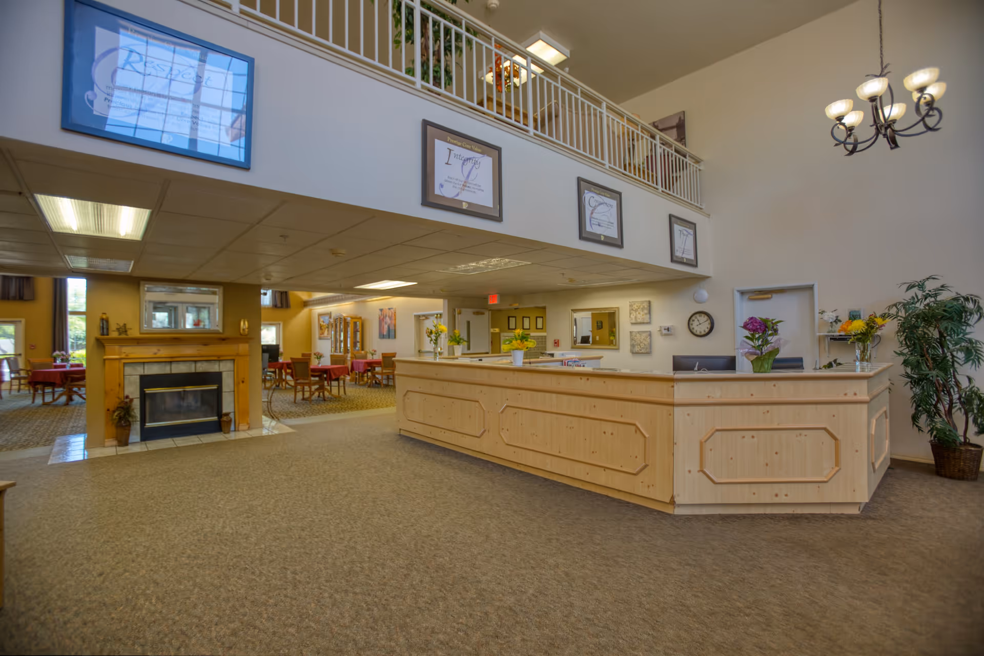 Interior view of a senior living facility reception area with a wooden front desk, a chandelier hanging from the ceiling, framed certificates on the wall above, a fireplace to the left, and a dining area with tables and chairs in the background.