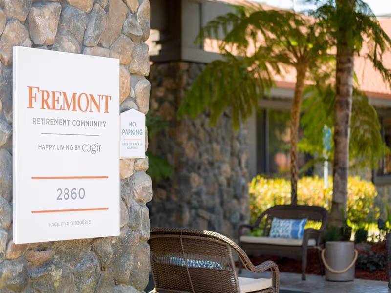 Outdoor seating area with wicker chairs and cushions near a stone wall with a sign that reads 'Fremont Retirement Community, Happy Living by Cogir, 2860'. There are palm trees and plants in the background.