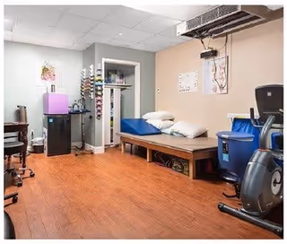 A therapy or rehabilitation room with exercise equipment including a stationary bike, a treatment table with pillows, a small refrigerator, and shelves with various supplies. The room has wood flooring and light-colored walls with anatomical posters.