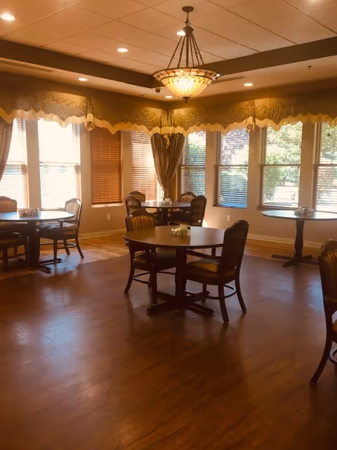 A cozy dining room with round wooden tables and cushioned chairs. Large windows with blinds and decorative valances allow natural light to fill the room. A chandelier hangs from the ceiling, illuminating the space with warm light.
