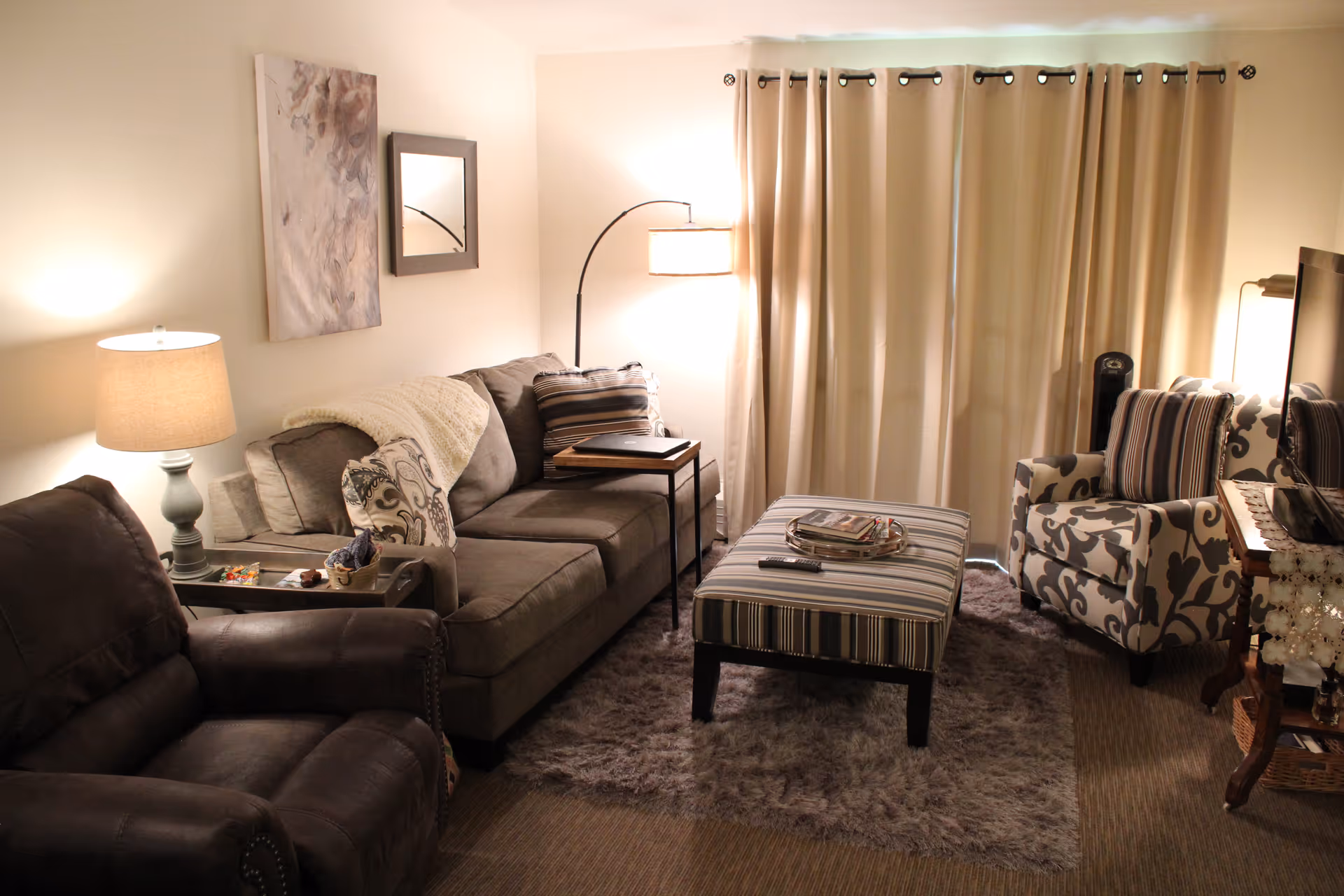 A cozy living room with a brown leather recliner, a gray fabric sofa with patterned pillows and a throw blanket, a striped ottoman with magazines and a remote control on top, a patterned armchair with a striped pillow, a floor lamp, a table lamp, a wall mirror, and a piece of abstract wall art. Beige curtains cover the window, and a soft rug is on the floor.