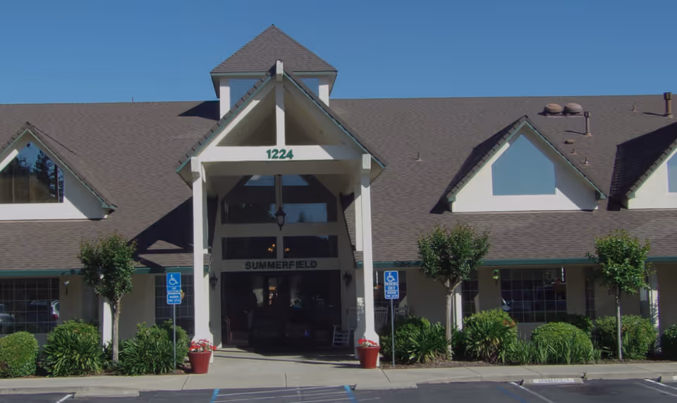 Front exterior view of Summerfield Senior Living facility with a peaked roof entrance, large windows, and landscaping including bushes and small trees. Handicap parking signs are visible near the entrance.