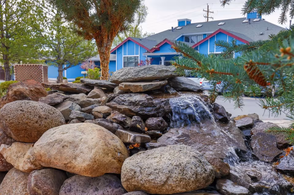 A stone water feature and rock landscaping in front of a blue building with trees and parked cars.