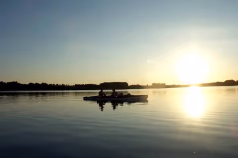 Two people paddling a canoe on a calm lake during sunset with the sun low on the horizon and trees in the distance.