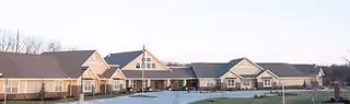 Exterior view of a single-story senior living facility building with multiple peaked roofs and large windows, set against a clear sky with a paved driveway and some landscaping in front.